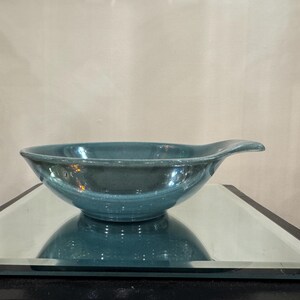 May include: A teal-colored ceramic bowl with a handle. The bowl has a smooth, glossy finish and is sitting on a mirrored surface. The bowl's shape is round with a slightly flared rim.