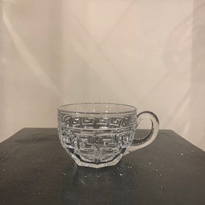 May include: Clear glass teacup with a handle. The cup features a geometric pattern around the rim and body. The cup is sitting on a dark surface.