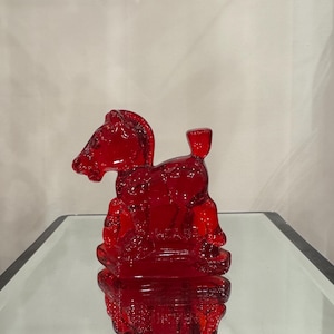 May include: A striking red glass horse figurine, featuring detailed craftsmanship. The horse is shown standing, complete with saddle and reins. The figurine is presented on a mirrored surface, accentuating its reflective properties.
