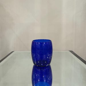 May include: A cobalt blue glass tumbler with a rounded shape. The glass is smooth and reflects light, sitting on a mirrored surface. The tumbler's vibrant color and simple design suggest a modern aesthetic.
