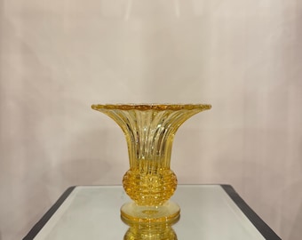 May include: A translucent amber-colored glass vase featuring a flared, scalloped rim and a ribbed neck that transitions into a textured, bulbous midsection. The vessel stands on a circular base, showcasing a clear, reflective quality throughout.