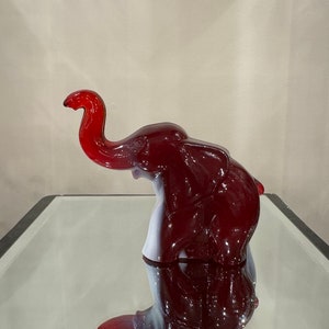 May include: A red glass elephant figurine with a raised trunk. The elephant is standing on a mirrored surface.