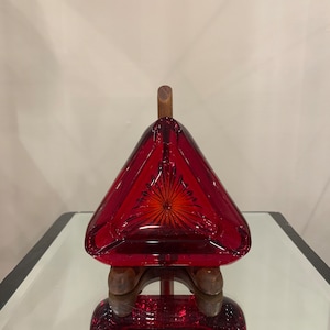 May include: A ruby red glass decanter with a triangular shape and a wooden stopper. The decanter has a starburst design on the front and sits on a wooden base. The glass is highly reflective.