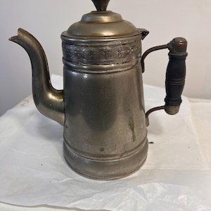 May include: Antique silver-toned metal coffee pot with a curved spout and a wooden handle. The lid is adorned with a decorative floral pattern and a small finial. The pot has a vintage aesthetic.