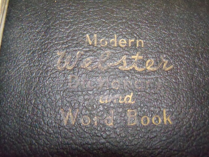May include: Close-up of a black leather-bound book with gold lettering that reads "Modern Webster Dictionary and Word Book".