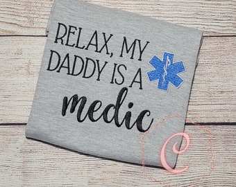 Può includere: Maglietta grigia con il testo "Relax, My Daddy Is A Medic" in lettere nere e una stella della vita ricamata in blu.