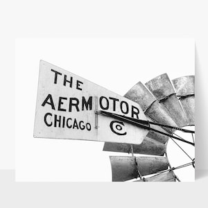 May include: Black and white photograph of a vintage windmill. The sign reads "THE AERMOTOR CHICAGO" in bold, black letters. The blades are made of metal and are partially visible. The image is set against a bright white background.