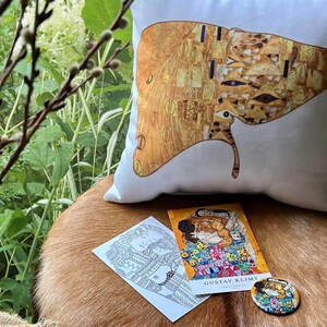 Puede incluir: Una almohada blanca con una impresión dorada y negra de El Beso de Gustav Klimt. La almohada está sobre una superficie de piel marrón con una impresión en blanco y negro de El Beso y un botón redondo con una imagen colorida de El Beso.