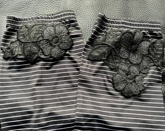 May include: A pair of black leggings featuring thin horizontal white stripes and an elastic waistband. Each leg is decorated with a large, black, shimmering floral lace appliqué with detailed leaf patterns stitched onto the upper thigh area.