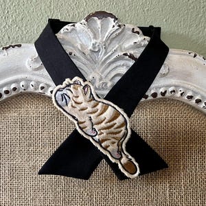 Pode incluir: Um choker de fita preta com um patch de gato bordado. O patch apresenta um gato dormindo com listras marrons e bege. O choker é exibido em uma moldura decorativa branca desgastada.