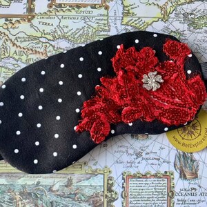 May include: A black sleep mask with white polka dots and a red floral design. The mask features a silver-coloured jewel in the centre of the floral design. It has a black elastic strap. The website www.BeExplores.net is visible.