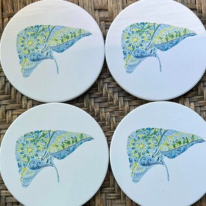Puede incluir: Cuatro posavasos de cerámica blanca con un diseño detallado y colorido de hígado. La ilustración del hígado presenta patrones florales azules, verdes y amarillos. Los posavasos están dispuestos sobre una superficie tejida marrón.