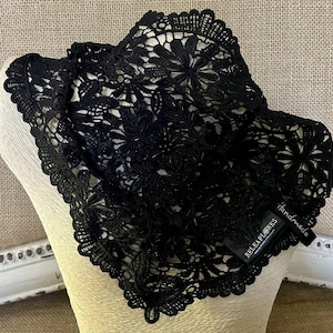 May include: A black lace head covering featuring an intricate floral pattern with scalloped edges. A small black fabric tag is attached to the side, displaying the white text BELEAPLORES and the word handmade in a cursive script font.