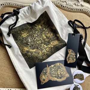 May include: A white tote bag with a black drawstring and a map design in gold and black. Also included are a black tag, a postcard, and two buttons, all featuring the same map design. The items are arranged on a neutral-toned surface.