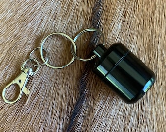 May include: A black, cylindrical keychain accessory with a silver-colored clasp and key rings. The accessory is made of a shiny, smooth material. The background is a textured, brown surface.