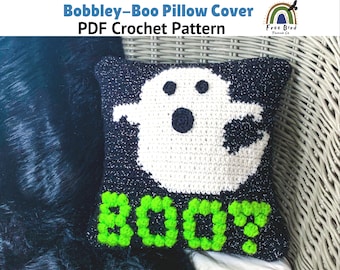 May include: A crocheted pillow cover featuring a white ghost with black eyes and a black mouth, with the word "BOO!" in green yarn below the ghost. The pillow cover is on a wicker chair.