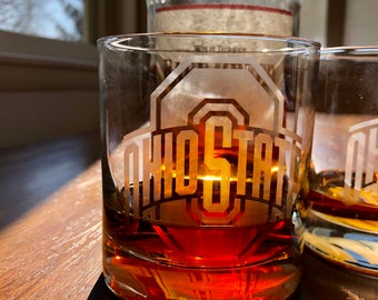 May include: Two clear glass tumblers with the words "Ohio State" etched on the front. The tumblers are filled with a dark amber liquid.