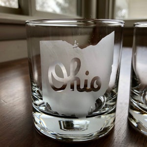 May include: Clear glass tumbler with the state of Ohio etched on the front. The word "Ohio" is etched within the state outline. The glass has a thick base and a straight-sided design. The glass is designed for drinking.