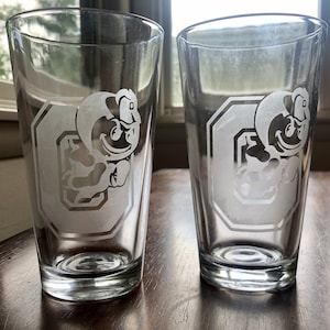 May include: Two clear glass pint glasses with an etched design of a cartoon mascot wearing a hat and holding a football. The mascot is inside a white outlined hexagon with the letter "O" inside.