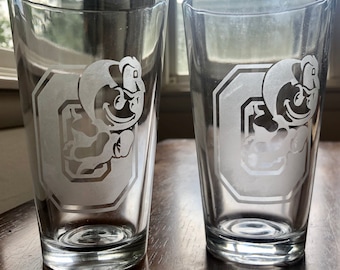 May include: Two clear glass pint glasses with an etched design of a cartoon mascot wearing a hat and holding a football. The mascot is inside a white outlined hexagon with the letter "O" inside.