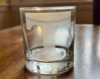 May include: A clear glass tumbler with a frosted white letter "T" etched on the front. The tumbler is sitting on a wooden surface.