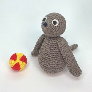 Puede incluir: Un juguete de foca de crochet gris con ojos blancos y nariz negra, sentado junto a una pelota de crochet roja y amarilla.