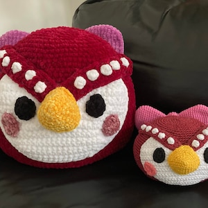 Puede incluir: Dos peluches de búho de crochet, uno grande y uno pequeño. El búho más grande es rojo con detalles blancos y un pico amarillo. El búho más pequeño es rosa con detalles blancos y un pico amarillo.