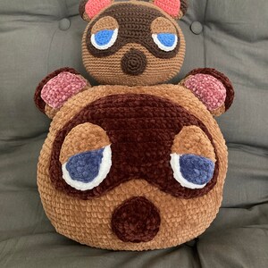 Op de afbeelding: Twee gehaakte pluche speelgoedjes van het Animal Crossing-personage Tom Nook. Het grotere pluche speelgoed is bruin met een bruine neus en blauwe ogen. Het kleinere pluche speelgoed zit bovenop het grotere pluche speelgoed en is bruin met een roze neus en blauwe ogen.