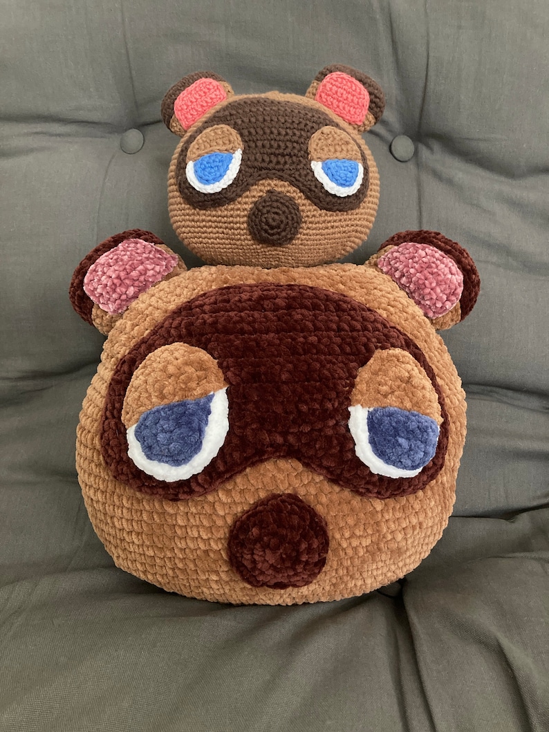 Op de afbeelding: Twee gehaakte pluche speelgoedjes van het Animal Crossing-personage Tom Nook. Het grotere pluche speelgoed is bruin met een bruine neus en blauwe ogen. Het kleinere pluche speelgoed zit bovenop het grotere pluche speelgoed en is bruin met een roze neus en blauwe ogen.