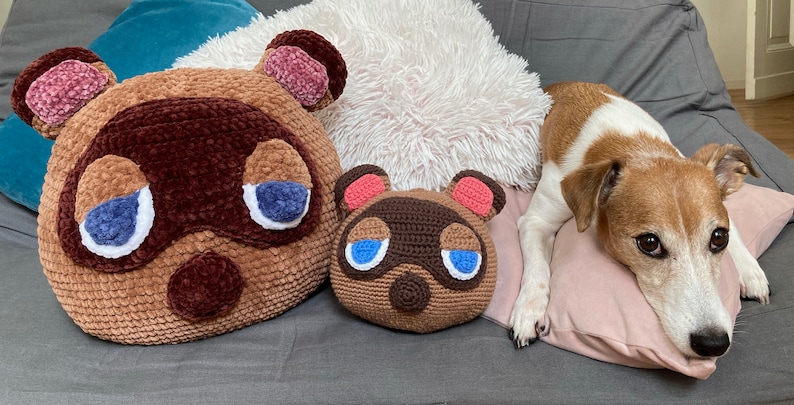 Op de afbeelding: Twee bruine gehaakte dierenknuffels met blauwe ogen en roze oren, een grotere en een kleinere, liggen op een grijze bank met een wit pluche kussen erachter. Een wit-bruine hond ligt op een roze kussen op de voorgrond.