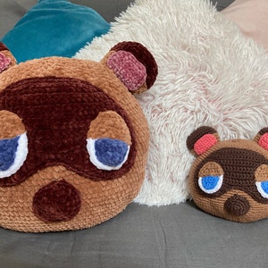 Op de afbeelding: Twee gehaakte pluche speelgoedjes van het Animal Crossing-personage Tom Nook. Het grotere pluche speelgoed is bruin met roze oren en blauwe ogen. Het kleinere pluche speelgoed is bruin met roze oren en blauwe ogen. Beide pluche speelgoedjes zitten op een grijze bank.