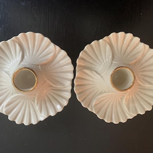 May include: Two white ceramic candle holders with gold accents. Each holder features a scalloped design with a central ring for a candle.