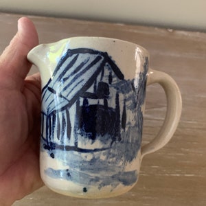 May include: A small, white ceramic creamer with a blue and white painted design of a barn. The creamer has a handle and a spout.