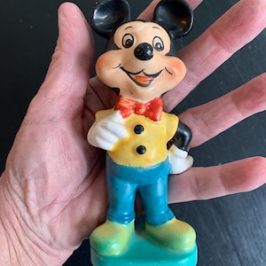 Peut inclure: Une figurine en c&eacute;ramique vintage de Mickey Mouse. Il porte une chemise jaune avec des boutons noirs, un pantalon bleu et un n&oelig;ud papillon rouge. Il a un grand sourire et se tient sur une base verte.