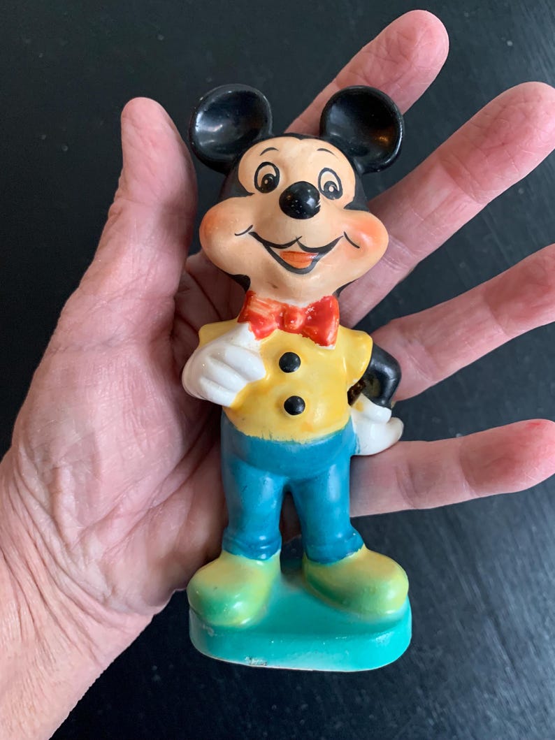 Peut inclure: Une figurine en c&eacute;ramique vintage de Mickey Mouse. Il porte une chemise jaune avec des boutons noirs, un pantalon bleu et un n&oelig;ud papillon rouge. Il a un grand sourire et se tient sur une base verte.