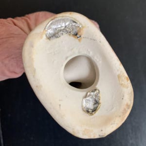 Peut inclure: Le dessous d'une figurine en c&eacute;ramique blanche avec un trou au centre. Il y a deux autocollants argent&eacute;s sur le fond de la figurine.