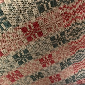 May include: A woven textile fabric featuring a repeating geometric pattern in shades of red, dark green, and beige. The material displays a textured surface with interlaced threads forming small square motifs arranged in a grid-like structure.