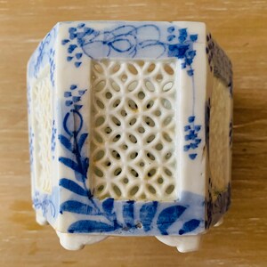 May include: A white ceramic planter with a blue floral design. The planter has a lattice-style cutout on the sides and is supported by small feet. The blue design includes flowers and vines.