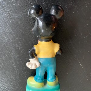 Peut inclure: Une figurine en c&eacute;ramique vintage de Mickey Mouse. La figurine est peinte en noir, jaune et bleu. Mickey porte une chemise jaune, un pantalon bleu et des gants blancs. Il est debout sur une base verte.