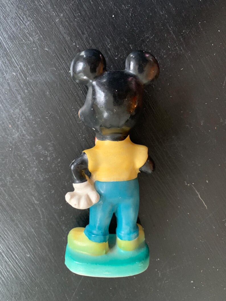 Peut inclure: Une figurine en c&eacute;ramique vintage de Mickey Mouse. La figurine est peinte en noir, jaune et bleu. Mickey porte une chemise jaune, un pantalon bleu et des gants blancs. Il est debout sur une base verte.
