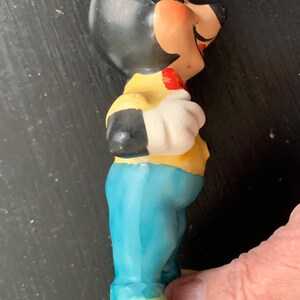 Peut inclure: Une figurine en c&eacute;ramique de Mickey Mouse portant une chemise jaune, un pantalon bleu et des chaussures rouges. Il est debout, la main droite sur la hanche et la main gauche tenant un objet rouge.