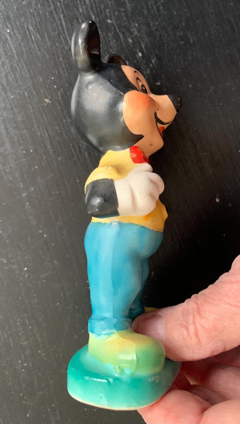 Peut inclure: Une figurine en c&eacute;ramique de Mickey Mouse portant une chemise jaune, un pantalon bleu et des chaussures rouges. Il est debout, la main droite sur la hanche et la main gauche tenant un objet rouge.