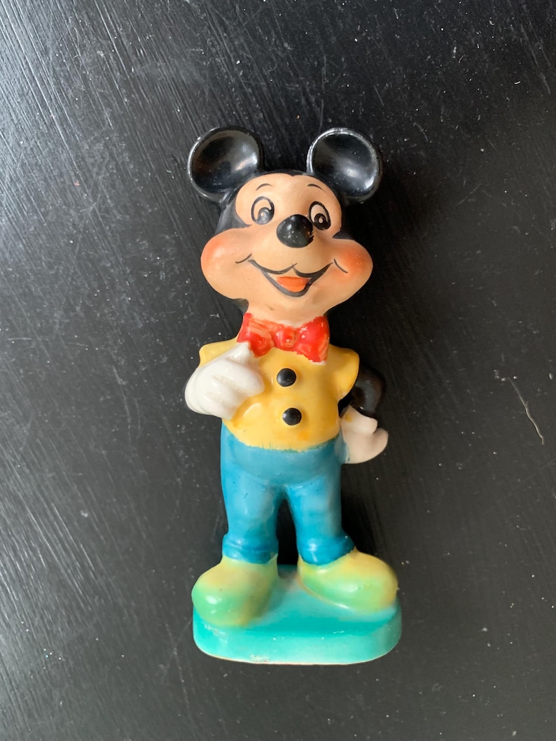 Peut inclure: Une figurine en c&eacute;ramique vintage de Mickey Mouse portant une chemise jaune avec des boutons noirs, un n&oelig;ud papillon rouge et un pantalon bleu. Il est debout sur une base bleue.