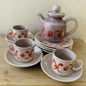 May include: A purple and white ceramic tea set with red floral designs. The set includes a teapot, four cups, and four saucers.