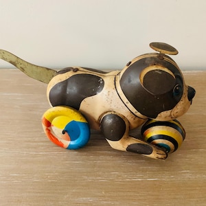 May include: Vintage wind-up toy dog with a cream and brown body, black spots, and a yellow, blue, and red wheel. The dog holds a striped ball and has a metal tail. A classic collectible toy.