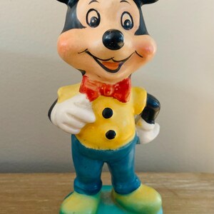Peut inclure: Une figurine en c&eacute;ramique vintage de Mickey Mouse portant une chemise jaune, un pantalon bleu et un n&oelig;ud papillon rouge. La figurine est debout sur une base bleue.