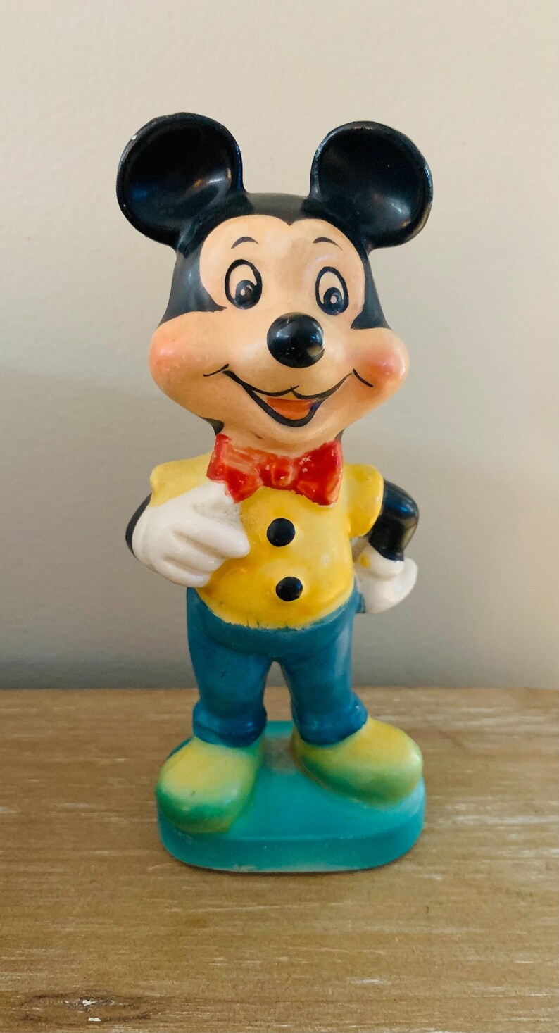 Peut inclure: Une figurine en c&eacute;ramique vintage de Mickey Mouse portant une chemise jaune, un pantalon bleu et un n&oelig;ud papillon rouge. La figurine est debout sur une base bleue.