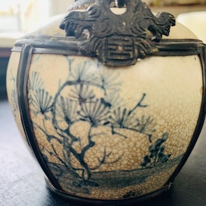May include: An antique, cream-colored ceramic container with a detailed blue and white painted landscape design. The container has a metal frame and a decorative metal top with an ornate design.
