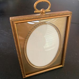 May include: A vintage gold-toned picture frame with an oval opening and a decorative handle. The frame has a rectangular shape with an ornate border and a glass front. The oval insert is white.