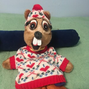 May include: A brown plush puppet with a white and red sweater, a blue hat, and brown pants. The puppet has a large mouth with white teeth and brown eyes.